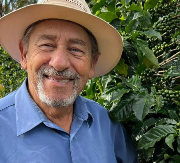 Man wearing a wide-brimmed hat and blue shirt standing in front of coffee plants with green beans.
