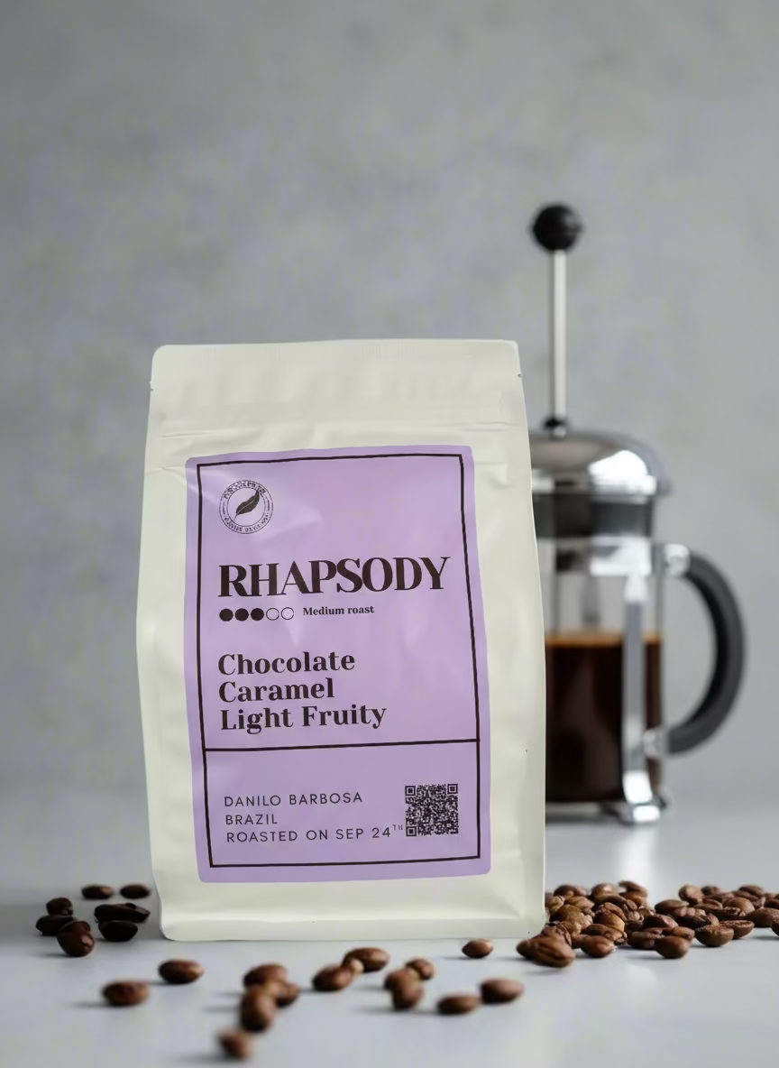 Rhapsody coffee package with a purple label on a gray background with coffee beans and a French press.