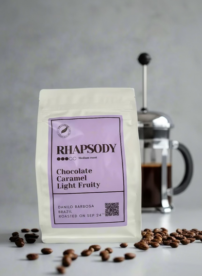 Rhapsody coffee package with a purple label on a gray background with coffee beans and a French press.